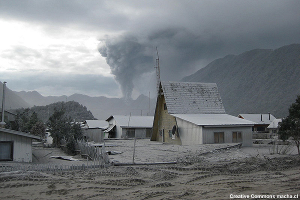 Houses buried in volcanic ash up to their first-floor windows in a town near the Chaiten volcano. The volcano plume is in the background.