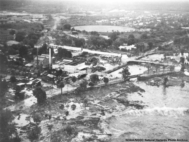 Hilo, Hawaii. Aerial view showing extent of innudation in area of Hilo Electric Company resulting from first wave as secondary wave approaches the area.